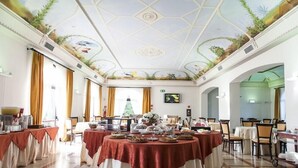 Un comedor amplio con varias mesas redondas cubiertas con manteles rojos, un mural grande en el techo y un árbol de Navidad al fondo.