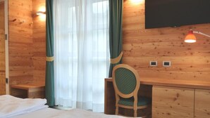 A wooden-paneled room with a bed, a desk, a chair, a TV, and a window with curtains. - Alp Holiday Dolomiti (Dimaro Folgarida)