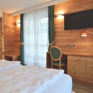 A wooden-paneled room with a bed, a desk, a chair, a TV, and a window with curtains. - Alp Holiday Dolomiti (Dimaro Folgarida)