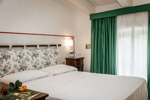 Ett hotellrum med en sänggavel dekorerad med blommor, ett nattduksbord med en lampa och ett fönster med gröna gardiner.