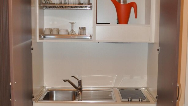 A compact kitchenette with a sink, dish rack, and a small cabinet with a red pot.