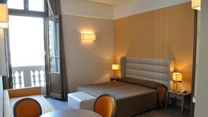 A hotel room with a bed, a small table, two chairs, a lamp, and a window with a view.