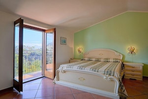 Un dormitorio con una cama grande, una mesita de noche, una mesita junto a la cama, un ventanal corredero que da a un balcón y un cuadro enmarcado en la pared.
