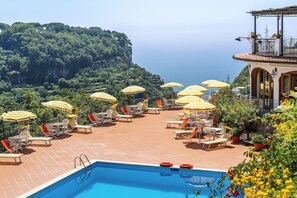 Poolbereich mit Liegestühlen und Sonnenschirmen, mit Blick auf eine Klippe und das Meer.