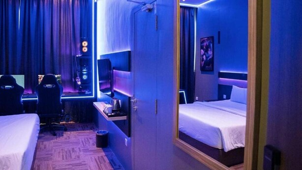 A hotel room with a bed, a desk with a computer and speakers, and a large mirror. - Sem9 Senai (Senai)