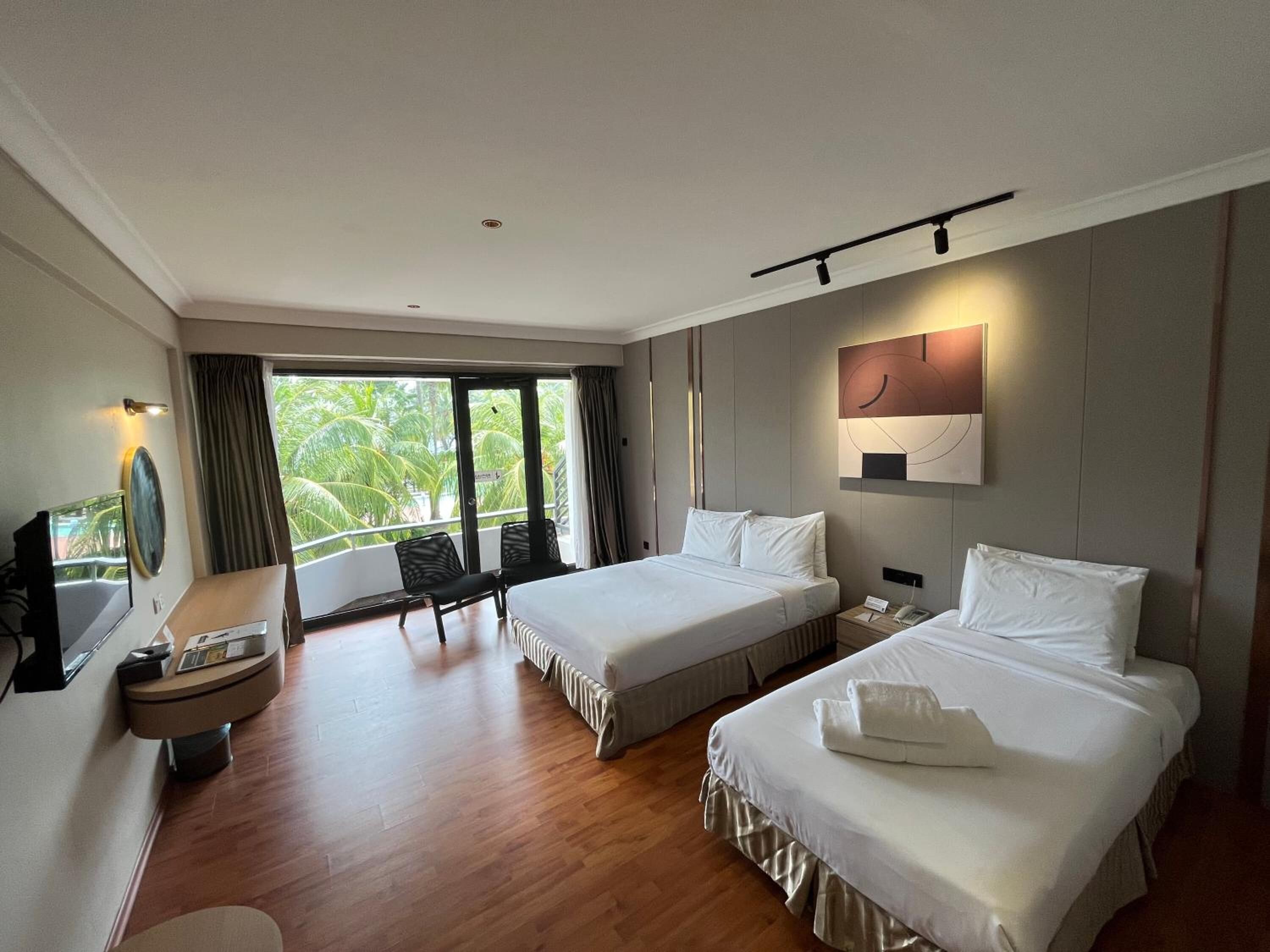 A hotel room with two beds, a desk with a TV, and a balcony with a view of palm trees.