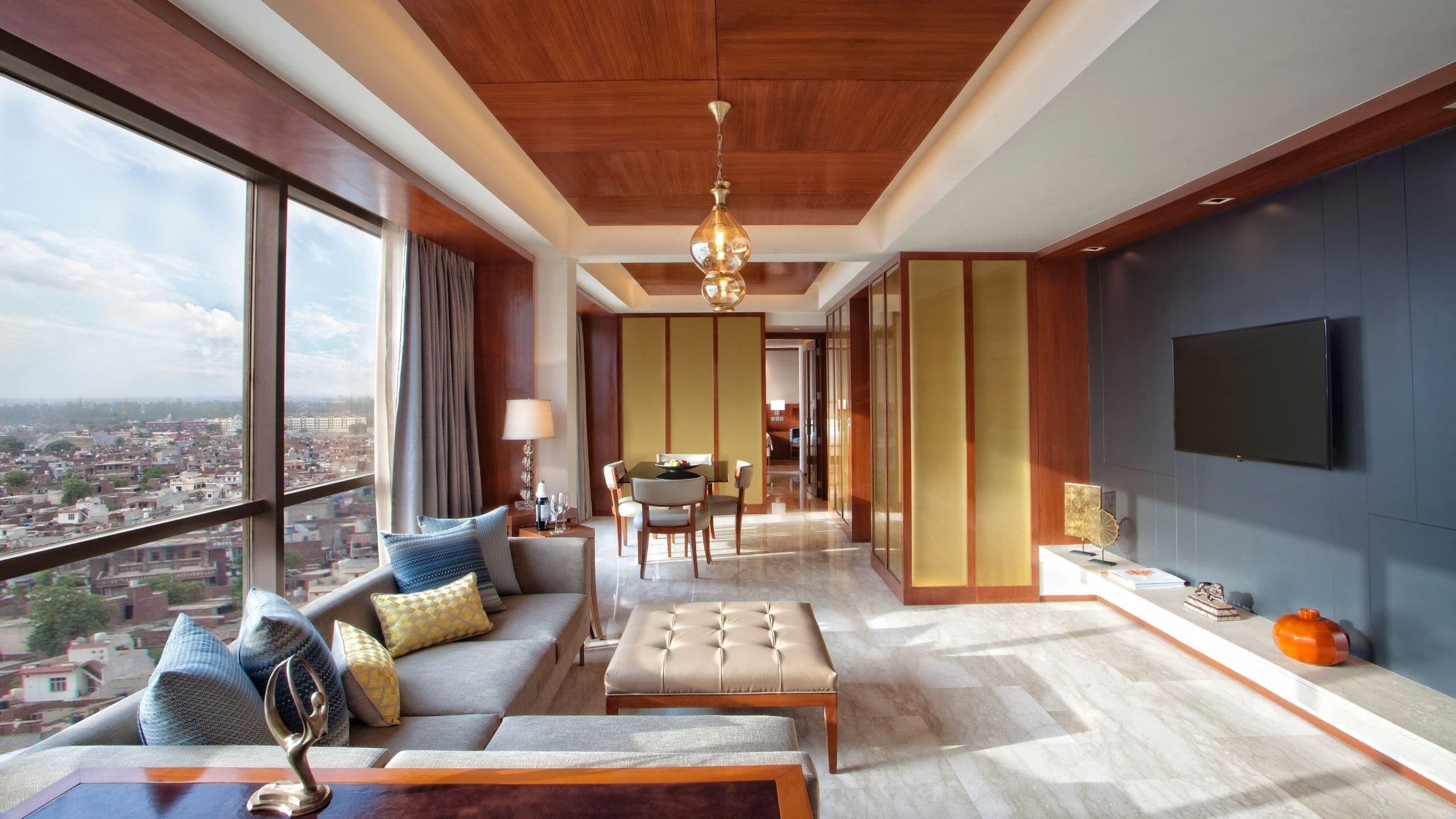 A modern hotel room with a large window offering a city view, a cozy seating area with a sofa and ottoman, a dining area with a table and chairs, and a flat-screen TV mounted on the wall.