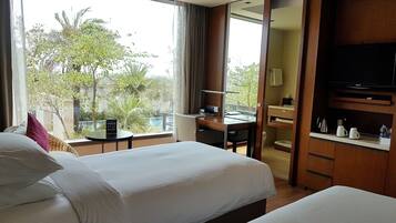 A hotel room with two beds, a desk, a TV, and a view of greenery outside.