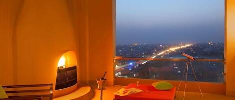 A room with a bed, a chair, a telescope, and a city view at dusk.