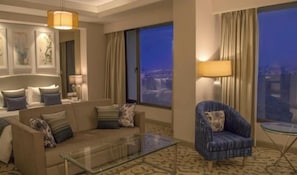 A hotel room with a sofa, armchair, and a glass coffee table. There are large windows with curtains, a bed with a headboard, and wall art.