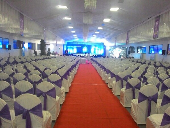 A large hall with rows of chairs covered in white and purple sashes, a red carpet, and a stage with a blue screen at the front. - Hotel Emerald Park- Sharanpur (Nashik)