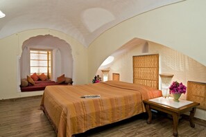 A bedroom with a large bed, a small table with a vase of flowers, and a window with patterned curtains. - Mandawa Castle (Jhunjhunu)