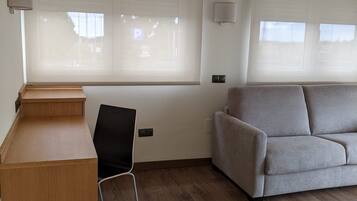 Una habitación con un escritorio de madera, una silla negra, un sofá gris y ventanas amplias con persianas.