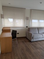 A room with a wooden desk, a black chair, a grey sofa, and large windows with blinds.