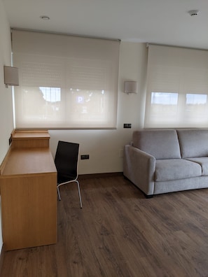 Una habitación con un escritorio de madera, una silla negra, un sofá gris y ventanas amplias con persianas.