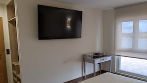 A modern bedroom with a large bed, a TV mounted on the wall, a small desk, and a window with blinds. - Cristal Aeropuerto (Castrillon)