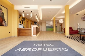Un vestíbulo de hotel con mostrador de recepción, un mapa mundial en la pared, un reloj y una alfombra azul con el texto 'HOTEL AEROPUERO'.