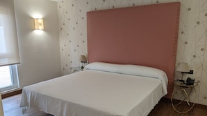 A large bed with a pink headboard, white bedding, and a small bedside table with a lamp. - Cristal Aeropuerto (Castrillon)