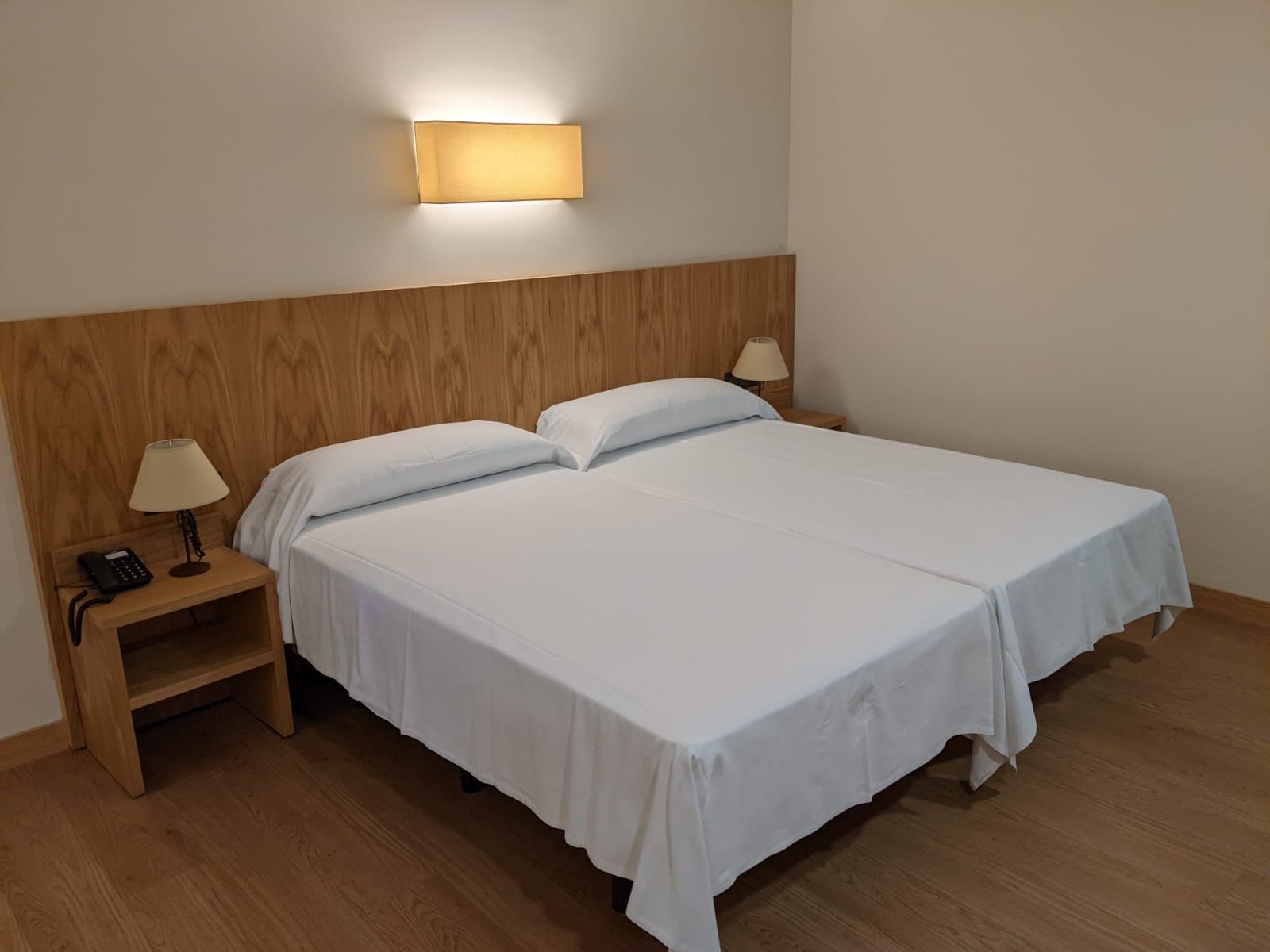 A hotel room with a wooden headboard, two beds with white linens, a bedside table with a lamp, and a wall-mounted light fixture.