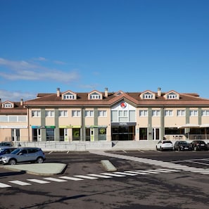 A hotel with a central entrance, multiple floors, and a parking lot with cars. - Cristal Aeropuerto (Castrillon)
