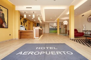 Um saguão de hotel com balcão de recepção, um mapa-múndi na parede, um relógio e um tapete azul com a inscrição 'HOTEL AEROPUerto'.