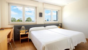 A hotel room with two single beds, a desk, a chair, a lamp, and a view of the outdoors through large windows. - Cristal Aeropuerto (Castrillon)