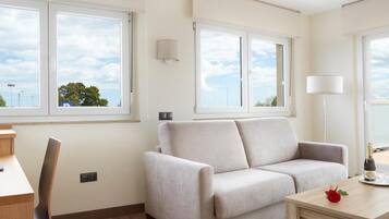 A modern living room with a sofa, a table, and a chair. There are large windows providing a view of the outdoors.