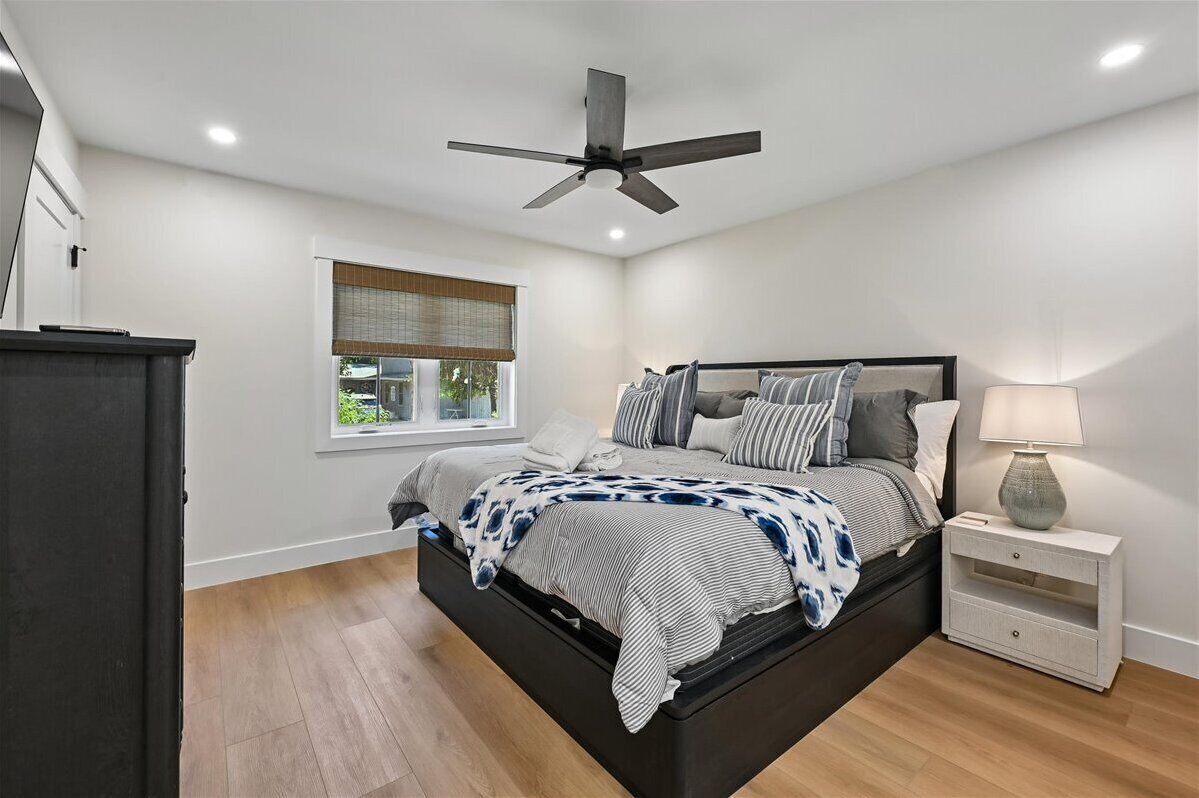 Quarto com cama, ventoinha no teto, mesa de cabeceira com abajur, janela com persianas e vista para o verde lá fora.