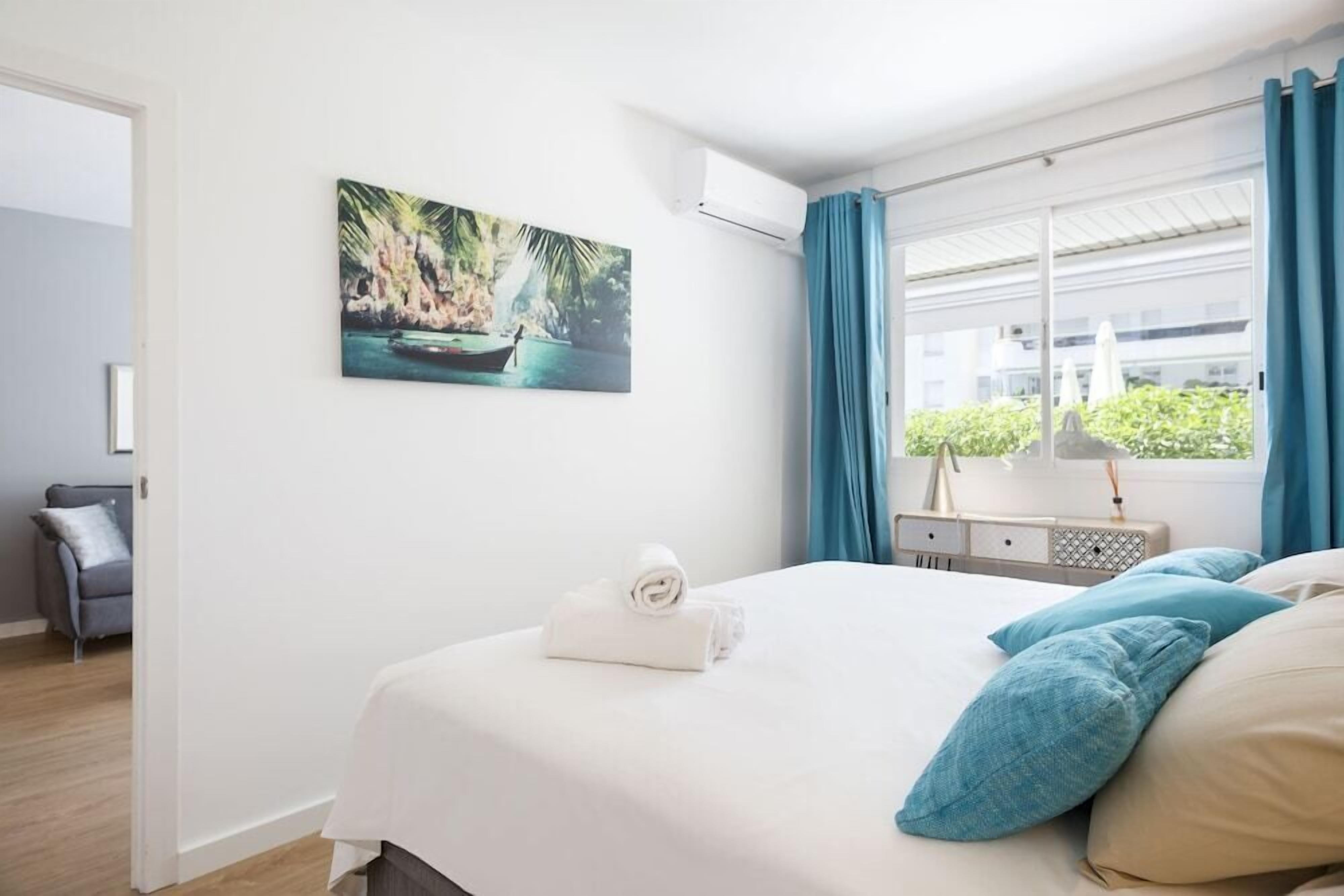 Una habitación de hotel con una cama, un cuadro de un paisaje tropical, una ventana con cortinas y un escritorio con una lámpara.