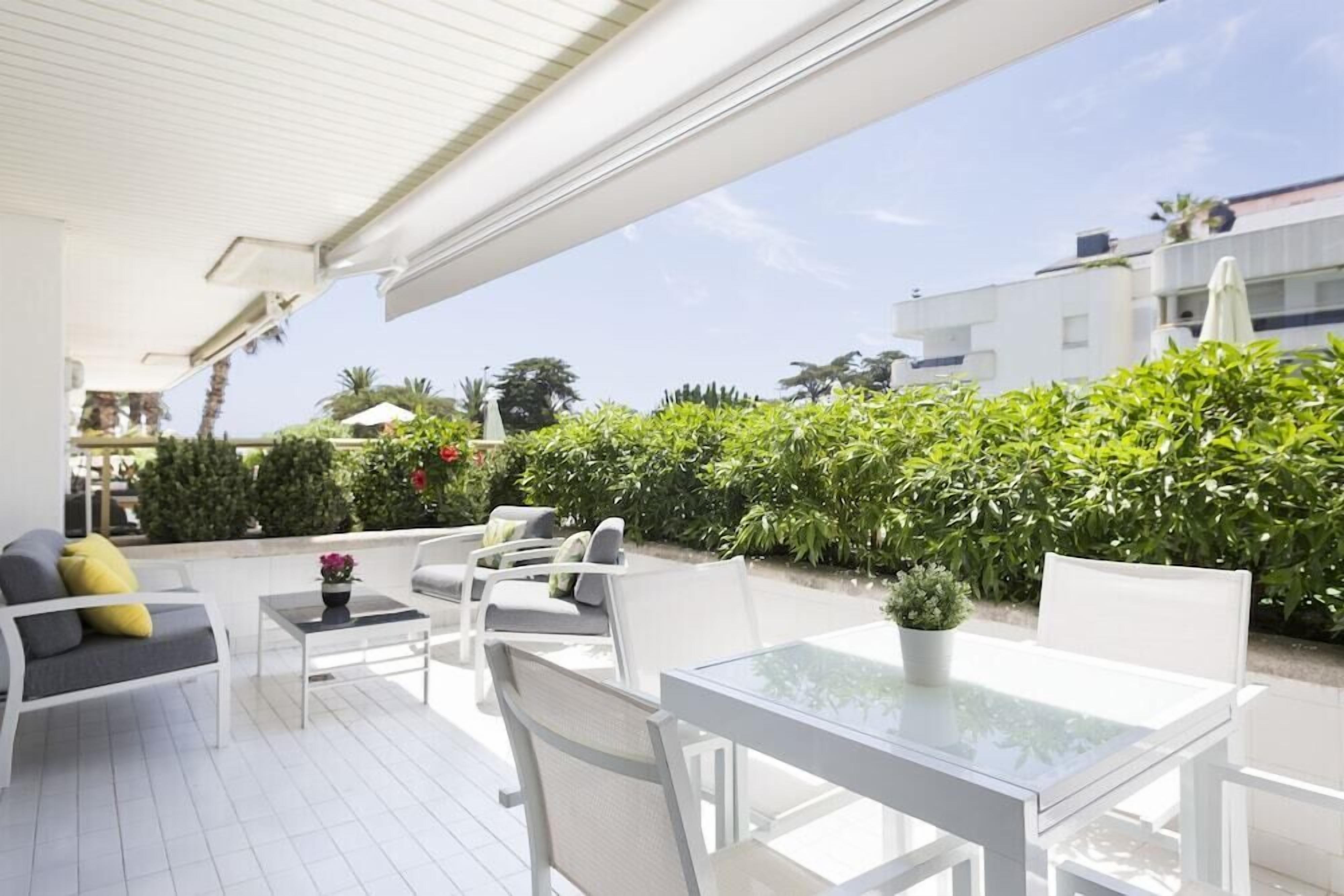 Un patio al aire libre con mobiliario blanco, incluyendo un sofá y sillas, una mesa de vidrio y una planta en maceta.
