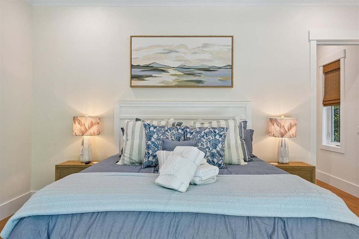 Un lit bien fait, recouvert d’une couvre-lit bleue, avec une tête de lit ornée d’oreillers à motifs, deux lampes de chevet et un tableau au-dessus du lit.