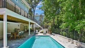 Ein Schwimmbad mit überdachter Terrasse, einer Treppe, die zu einer zweiten Etage führt, und einem von einer Hecke umgebenen Garten.