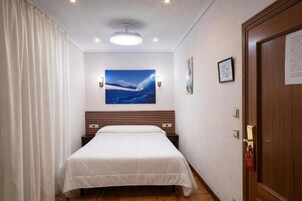 A hotel room with a bed, a ceiling light, a painting, and a door.