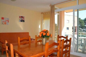 Un comedor con una mesa y sillas de madera, un jarrón con flores naranjas y vistas a un balcón exterior a través de un ventanal.