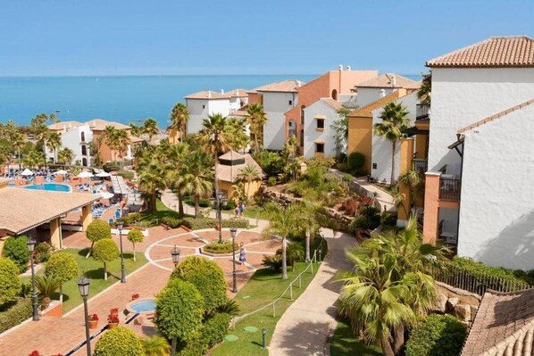 Un resort costero con zona de piscina, palmeras y edificios de estilo mediterráneo.