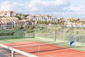 Una cancha de tenis con red, rodeada por una valla, y edificios residenciales al fondo.
