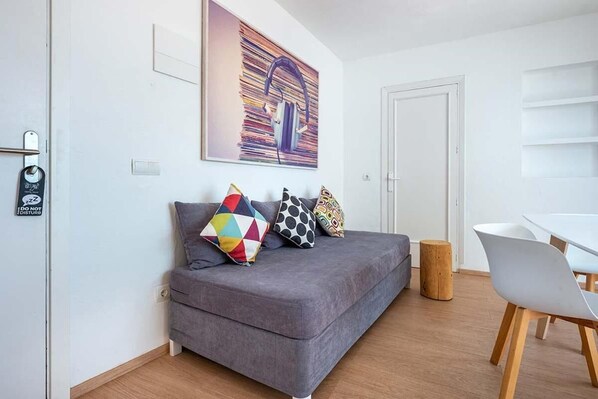 A modern living room with a grey sofa, colorful pillows, a wooden stool, and a white dining table with chairs. - Cenit Apartments (Ibiza Town)