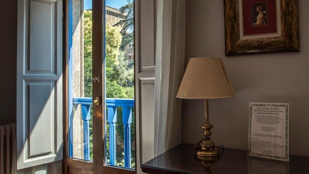 A room with a wooden door, a blue balcony railing, a table with a lamp, and a framed picture on the wall. - Don Juan de Austria (Jarandilla de la Vera)