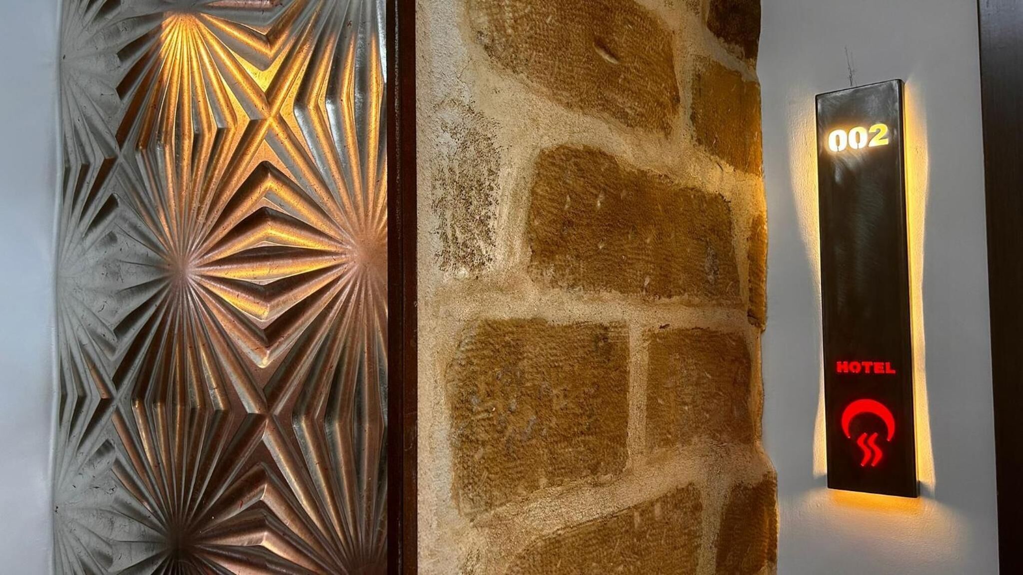 A wall with a geometric metal panel design and a stone wall with a digital display showing room number 002 and a red hotel icon.