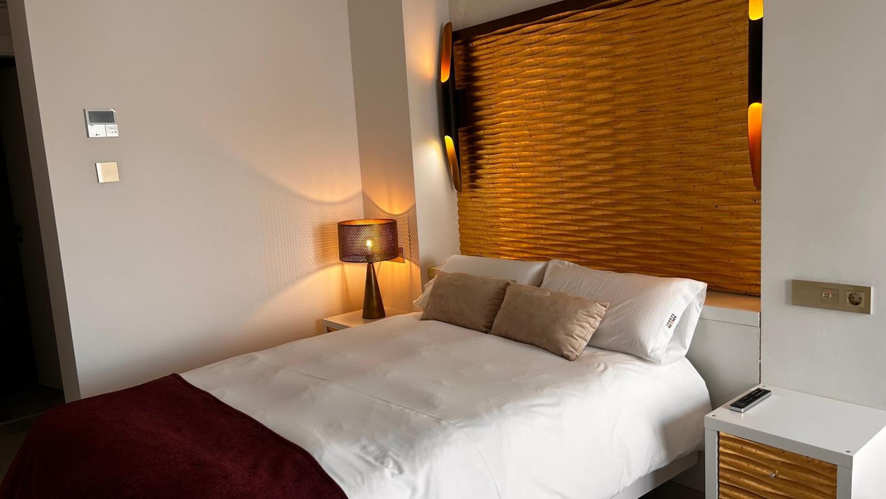 Ein Hotelzimmer mit Bett, Nachttisch, Lampe, Fenster mit Jalousien und einer Steckdose.