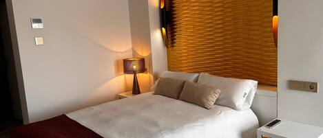 Ein Hotelzimmer mit Bett, Nachttisch, Lampe, Fenster mit Jalousien und einer Steckdose.