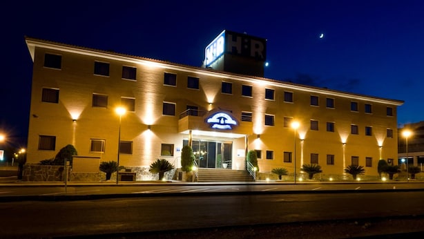 A multi-story hotel with a prominent 'HR' sign on the roof, a well-lit entrance, and a clear view of the street. - Complejo Hostelero Torrijos (Cheste)