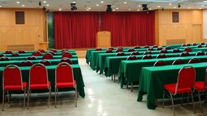 Ein Konferenzraum mit Tischen und Stühlen in Reihen, eine Bühne mit rotem Vorhang, einem Rednerpult und einer Dachkuppel.
