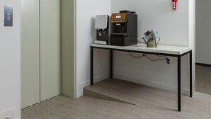 Un ascensor moderno y limpio con una pantalla digital, una máquina de café sobre una mesa y un detector de incendios en la pared.