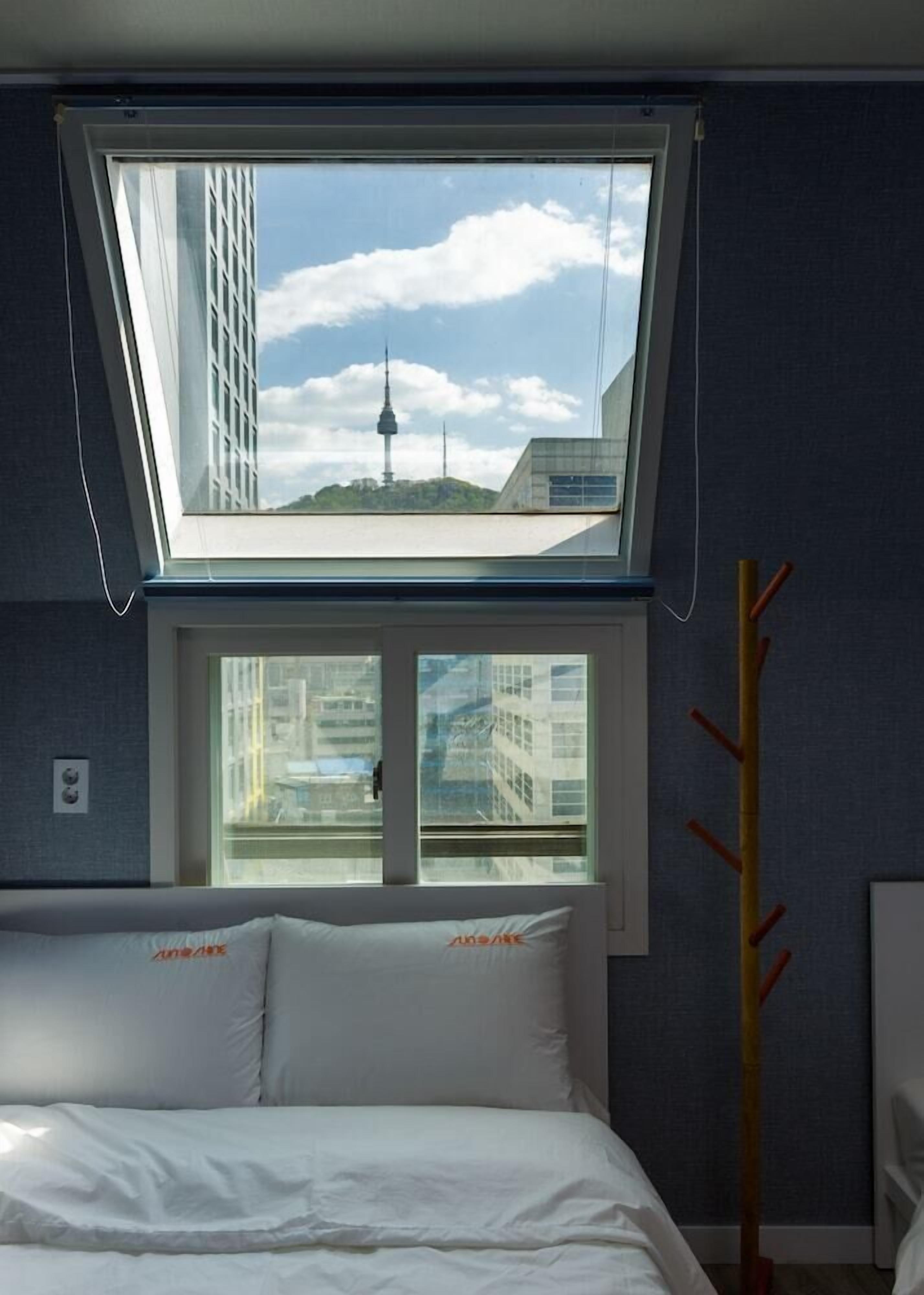 A bedroom with a skylight, a bed, a coat rack, and a view of a cityscape including a tower.