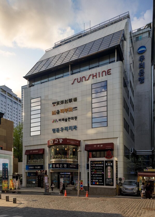 A modern building with the name 'SUNSHINE' prominently displayed. It features a large glass facade, solar panels on the roof, and multiple commercial spaces on the ground floor. - Myeongdong Sunshine Guesthouse (Seoul)