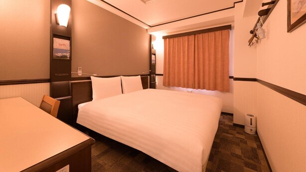 Habitación de hotel con cama, escritorio, silla, mesita de noche, lámpara, ventana con cortinas y un cuadro colgado en la pared.