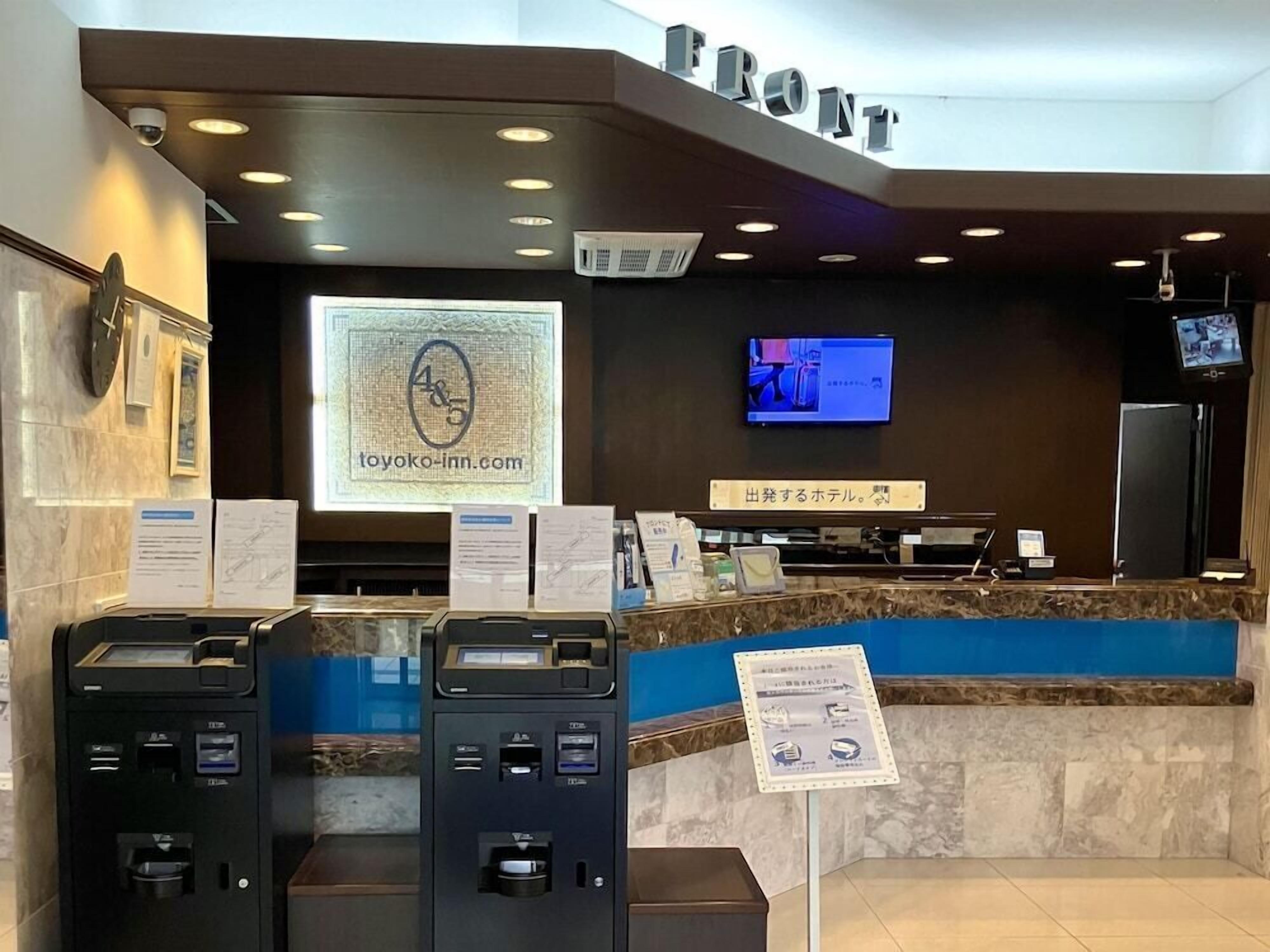 A hotel reception area with a marble counter, a digital screen displaying the hotel's website, a television, and two self-service kiosks.