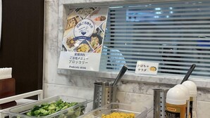 Zona de buffet con recipientes de comida etiquetados, un cartel anunciando productos locales y un dispensador de bebidas.