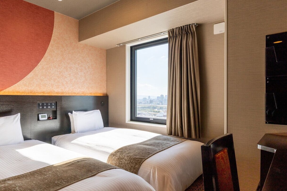 Une chambre d’hôtel avec deux lits, une grande fenêtre avec des rideaux, un téléviseur à écran plat et une vue sur la ville.